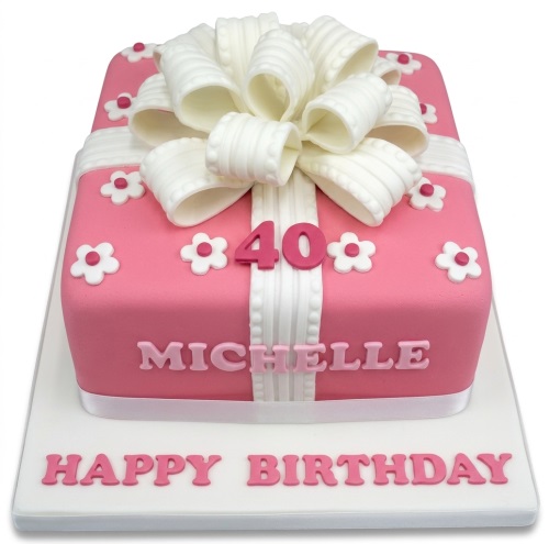 Pink gift box shaped birthday cake with white bow, ribbon, and small white flowers, with custom age and name.