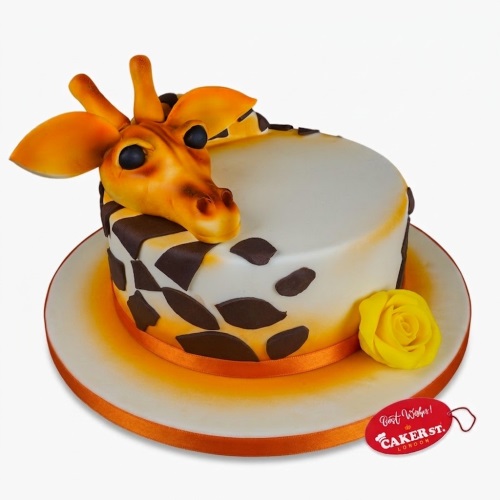 Round white cake with brown giraffe spots, a 3D giraffe head topper, orange ribbon, and a yellow sugar rose on the board.