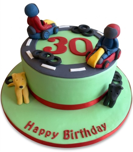 Two-tier green go-kart racing birthday cake with red custom age topper, drivers, tyres, road track, and 'Happy Birthday' message.