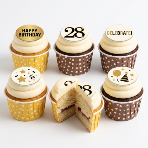Six cupcakes with cream frosting, some in yellow polka dot wrappers and some in brown. Toppers include 'Happy Birthday', '28', 'Celebrate!', stars, confetti, and a party hat with balloon. One cupcake is cut open revealing a jam filling.