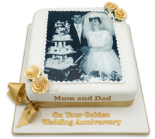 White anniversary cake with edible photo of a wedding couple, golden roses, and gold ribbon.