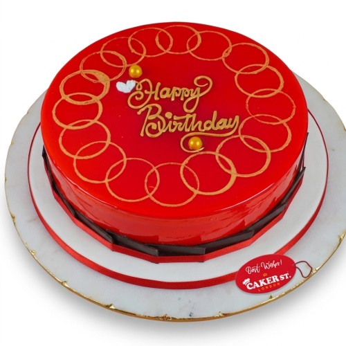 Round red glossy birthday cake with gold 'Happy Birthday' text and interlocking circle pattern, decorated with chocolate shards and a red ribbon.