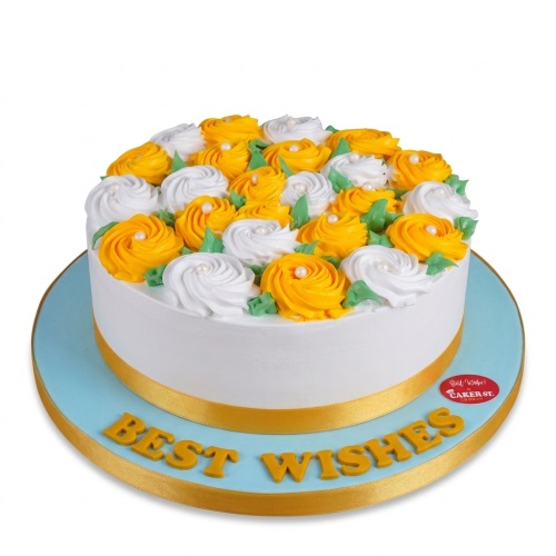 Round white cake with yellow and white frosting rosettes, green leaves, and gold 'Best Wishes' message.