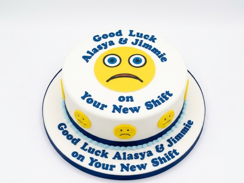 Round white fondant cake with a large sad emoji, smaller emojis, and blue 'Good Luck' message.
