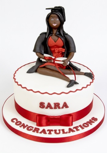 White single-tier cake with a female graduate figure on top, holding a diploma, with red ribbon and custom message.