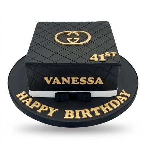Black quilted square cake with gold GG logo, gold 'Happy Birthday' message, white ribbon, and black bow.