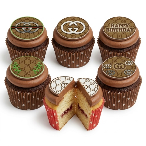 A selection of six chocolate cupcakes with brown frosting and round designer-style Gucci logo toppers, one saying 'Happy Birthday'. One cupcake is cut open revealing a light sponge and jam filling.