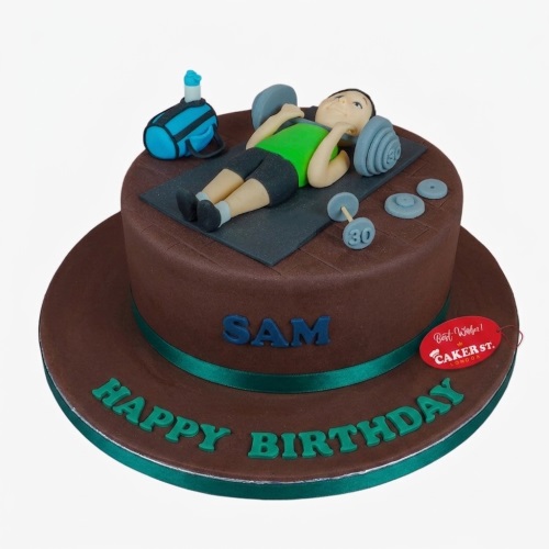 Round brown cake with a fondant figure of a man lifting weights, gym bag, water bottle, dumbbells, and personalised message.