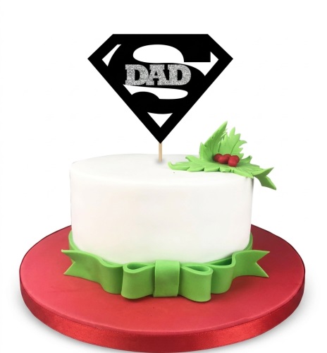 White single-tier cake with a black Superman-style 'DAD' topper, green ribbon, and holly leaves on a red board.
