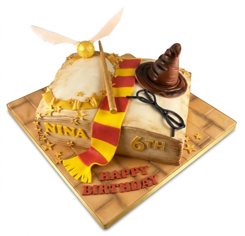 Book-shaped Harry Potter theme cake with golden snitch, wand, sorting hat, glasses, and red and yellow scarf.