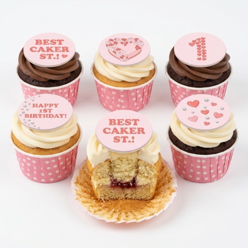 Pink 1st birthday cupcakes with heart toppers, 'Happy 1st Birthday' message, and polka dot wrappers.