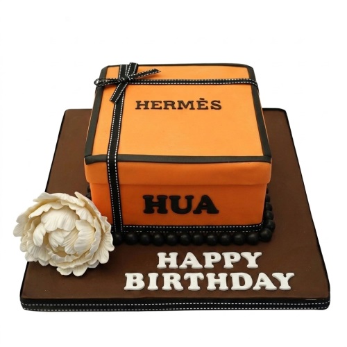 Orange Hermes style gift box cake with black ribbon, white flower, and 'Happy Birthday' message on a brown board.
