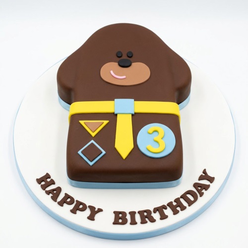 Hey Duggee character birthday cake in brown with yellow tie and custom age badge on a white board.