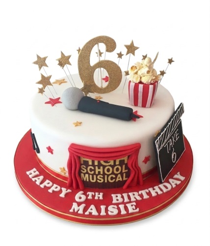 Round white High School Musical themed birthday cake with gold '6' topper, microphone, popcorn, and clapperboard.