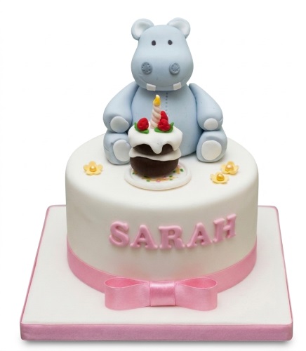 White round birthday cake with a blue hippo figure, a small cupcake with a candle, and a pink ribbon. Features a personalised name option.