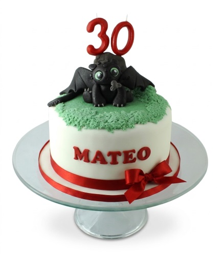 White round birthday cake with black Toothless dragon figure, green grass, red ribbon, and custom age candles.