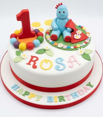 White round cake with Igglepiggle figurine, red number one topper, multicoloured balls, and personalised name.