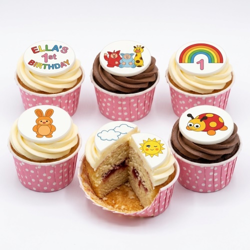 Assortment of 1st birthday cupcakes with animal, rainbow, sun, and character toppers in pink polka dot cases.