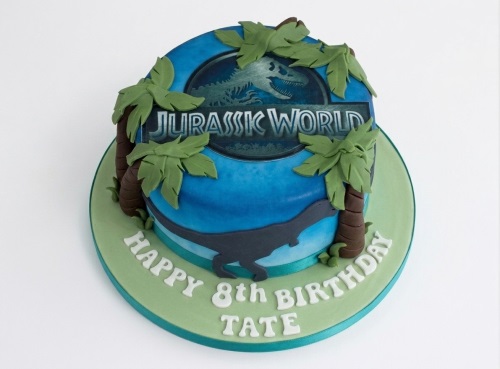 Round blue and green Jurassic World themed birthday cake with dinosaur and palm trees.