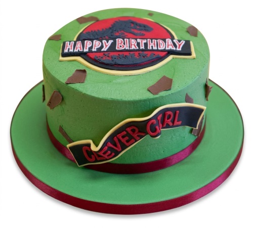 Round green Jurassic Park themed birthday cake with dinosaur logo and "Happy Birthday" message.