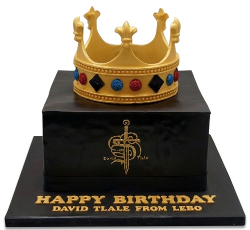 Black square birthday cake with a gold crown topper and gold sword emblem.