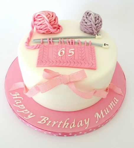 Round white birthday cake with pink and lilac yarn balls, knitting needles, and a pink knitted swatch with a custom age. Pink ribbon around the base.
