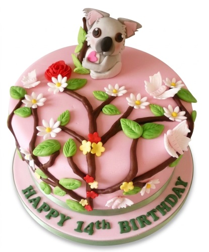 Pink birthday cake with 3D koala topper, branches, green leaves, white flowers, and butterflies.