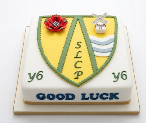 White square cake with a yellow and green school crest, poppy, windmill, and "GOOD LUCK" message.