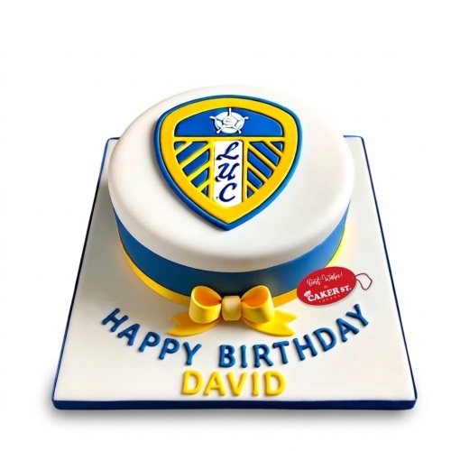Round white cake with Leeds United football club crest, blue and yellow ribbon, and a yellow bow. Customisable 'Happy Birthday' message.