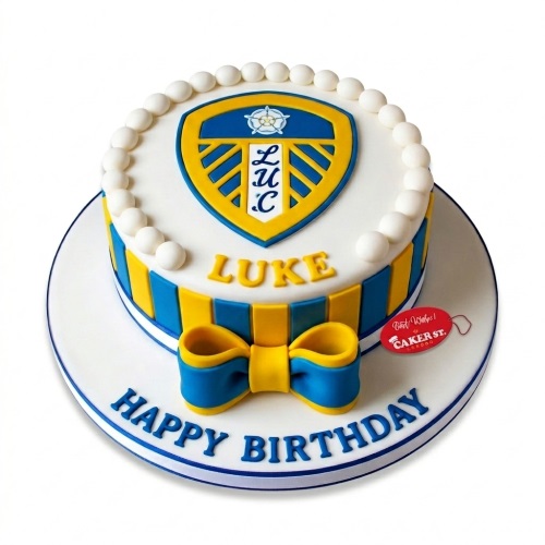 Round white birthday cake with Leeds United crest, blue and yellow stripes, and a bow. Personalised with a name and 'Happy Birthday'.