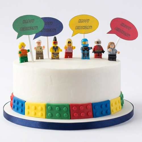 White birthday cake decorated with colourful building blocks around the base and various minifigure toppers holding 'Happy Birthday' speech bubbles.
