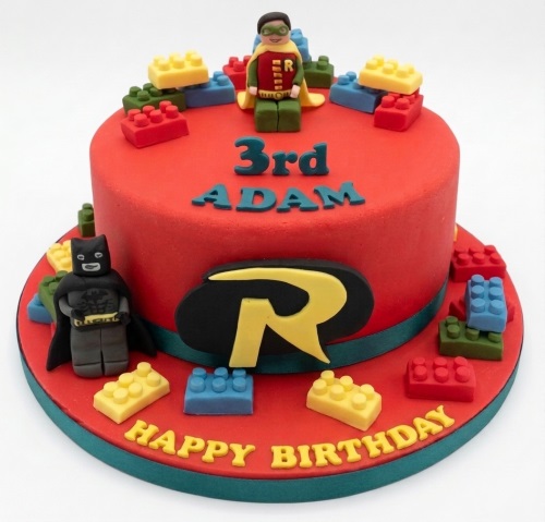 Red round birthday cake with Batman and Robin figures, yellow, red, blue, green building blocks, and a custom age and name.