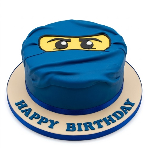 Round blue fondant cake decorated as a Lego Ninjago blue ninja face with yellow eye mask and black eyes, on a white cake board with 'Happy Birthday' message.