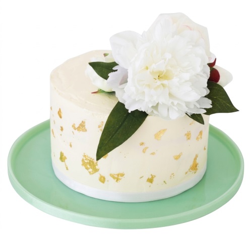 Elegant round cake with white peony flowers, green leaves, and gold leaf flakes on a light green board.