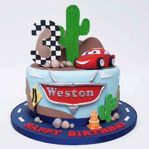 Single-tier racing car themed birthday cake with a red car, green cactus, checkered flag number topper, and road details.