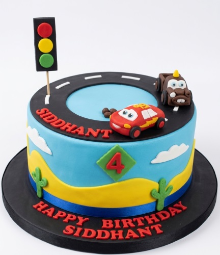 Round blue and yellow birthday cake with a black road, red race car, brown tow truck, traffic light, cacti, and a green custom age topper.