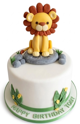 White round birthday cake with a 3D fondant lion topper, green leaves, and rocks.