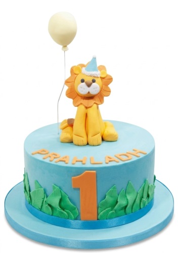 Blue round cake with a fondant lion, balloon, green leaves, and customisable age and name.