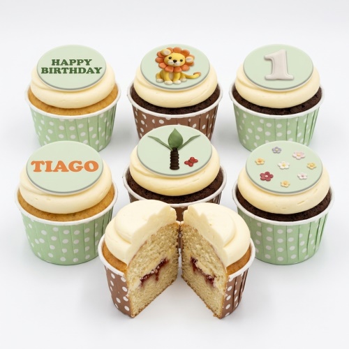 Assorted jungle safari themed cupcakes with cream frosting and edible toppers, including a lion, palm tree, flowers, and custom age '1' and name 'Tiago'.