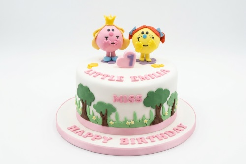 White round cake with Mr. Men Little Miss characters, green trees, grass, and a pink number one topper.