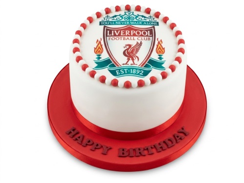 Round white fondant cake with Liverpool Football Club crest on top, red and white edible pearls, and a red ribbon. The cake board is red with 'Happy Birthday' written on it.