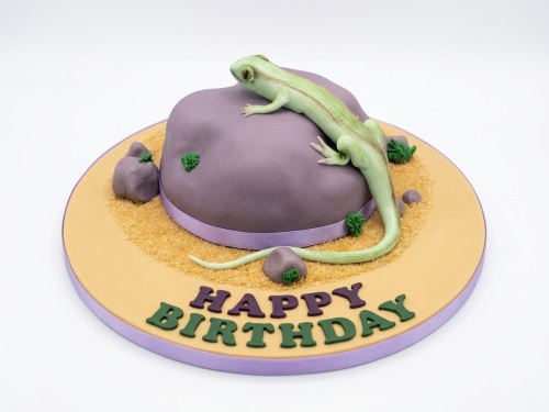 Green lizard figurine on a purple rock-shaped birthday cake with edible sand and "Happy Birthday" message.