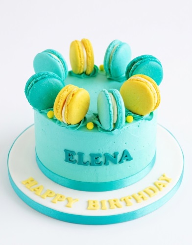 Round teal birthday cake topped with yellow and teal macarons and small yellow sprinkles, with a custom name on the side and a 'Happy Birthday' message on the board.