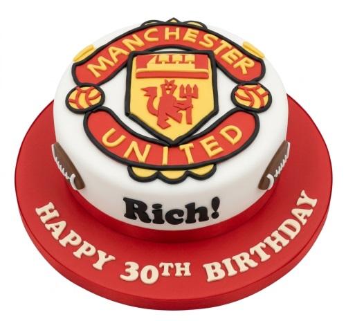 Round Manchester United themed birthday cake with club crest and football decorations on a red board with a custom message.