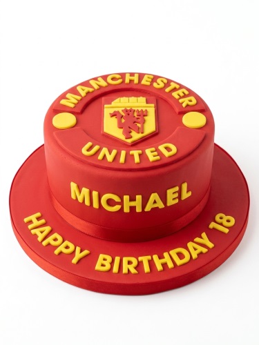 Red two-tier Manchester United themed birthday cake with yellow club logo and custom message.