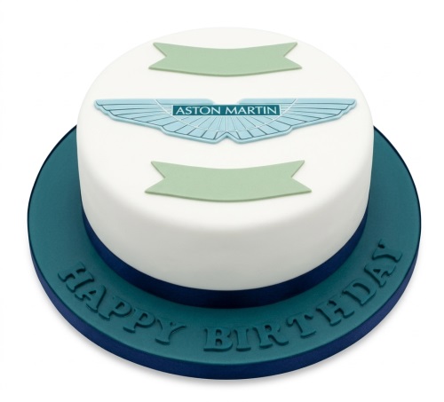 Round white Aston Martin logo birthday cake with green banners and 'Happy Birthday' on blue board.