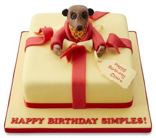 Square yellow gift box cake with red ribbon and an edible meerkat figure.
