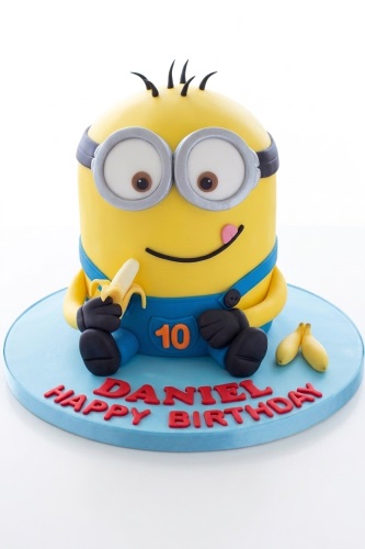 Yellow Minion character birthday cake with goggles, blue overalls, and a banana.