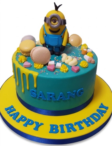 Round teal Minion birthday cake with yellow drip, macarons, sweets, and a 3D Minion topper.