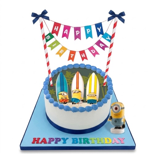 Round white birthday cake with Minions and surfboards, blue icing, and a 'Happy Birthday' banner and message.